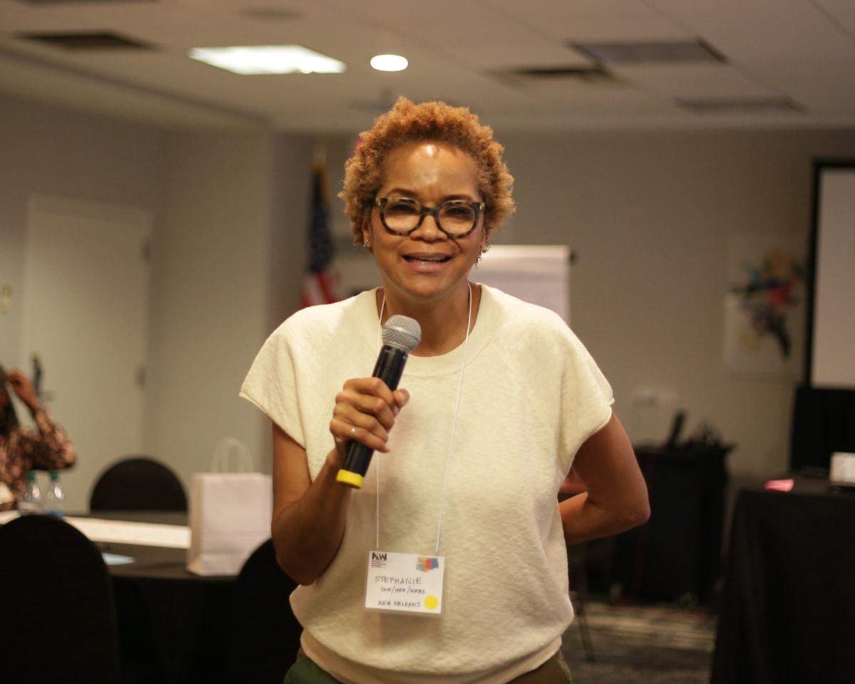 Stephanie Atkins, a Black woman, stands in a conference room holding a wireless microphone in her right hand, and smiles as she addresses an unseen audience. The photo is taken from the audience's point of view, so Stephanie appears to be smiling at and addressing the camera. Hanging from her neck is a name badge on a long white string.
