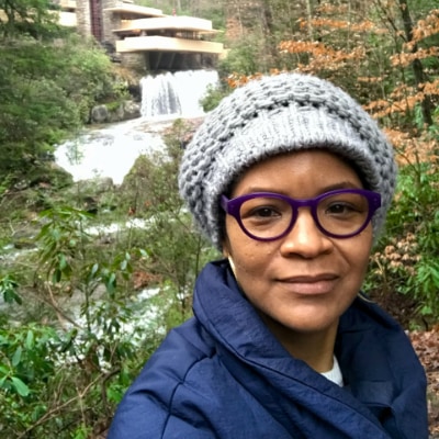 Stephanie Atkins, a Black woman, poses for a selfie with the Frank Lloyd Wright building "Falling Water" in background.