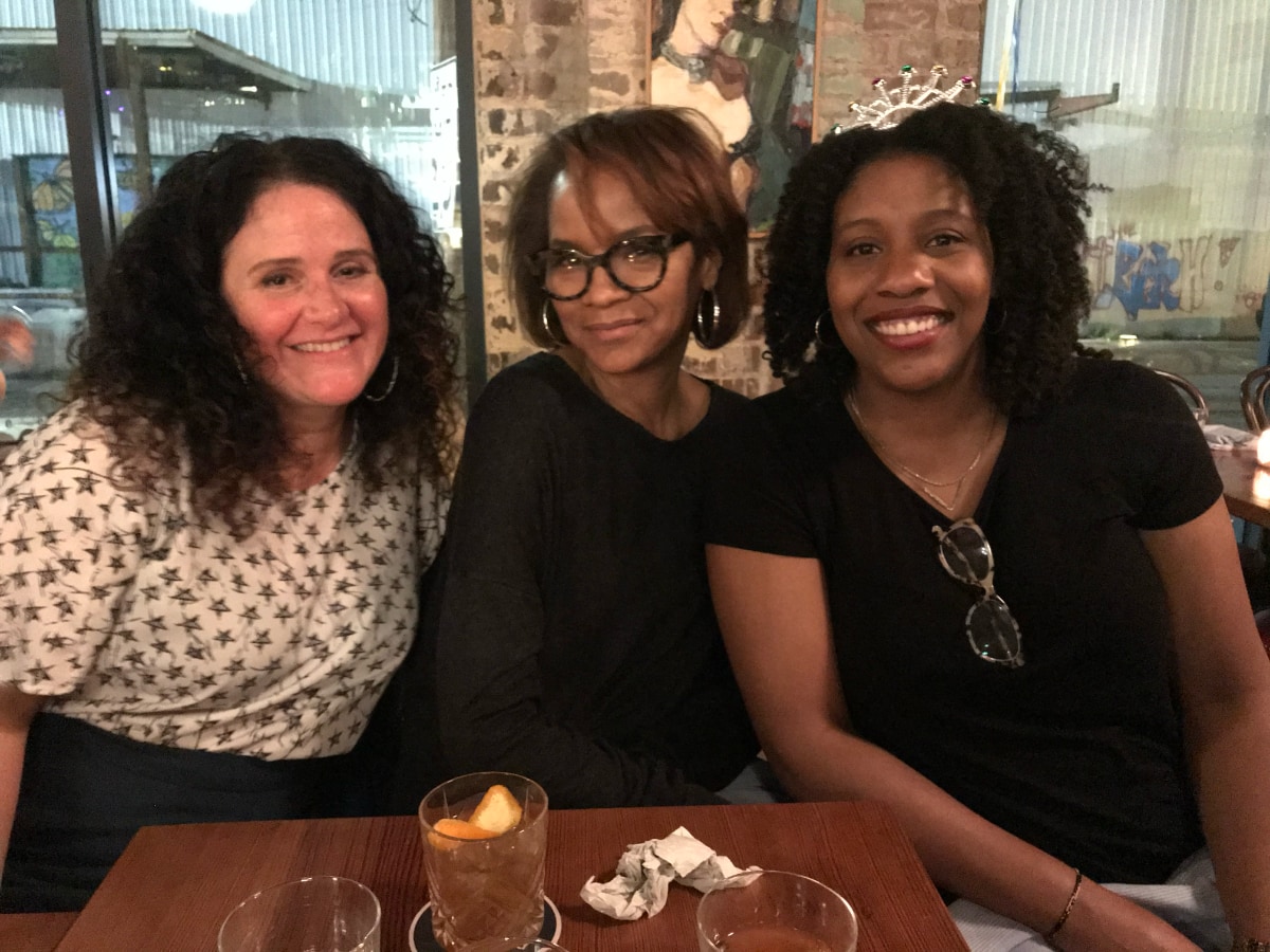 Stephanie poses with two other women at a table in a restaurant. On the left side of the photo is a white woman with dark shoulder-length hair. In the center is Stephanie Atkins, a Black woman. On the right is a Black woman with black hair almost to the shoulders.