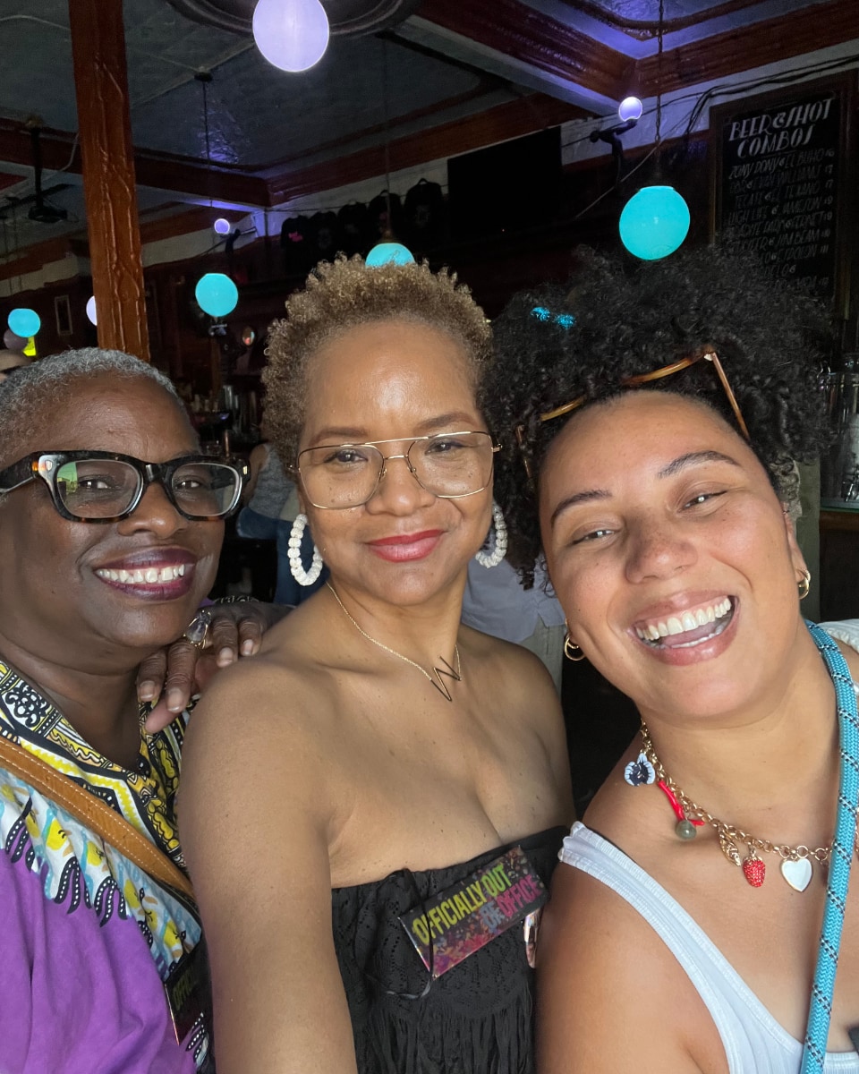 Stephanie Atkins, a Black woman, stands between two other women of color as the three smile for a group selfie in a restaurant during the daytime.
