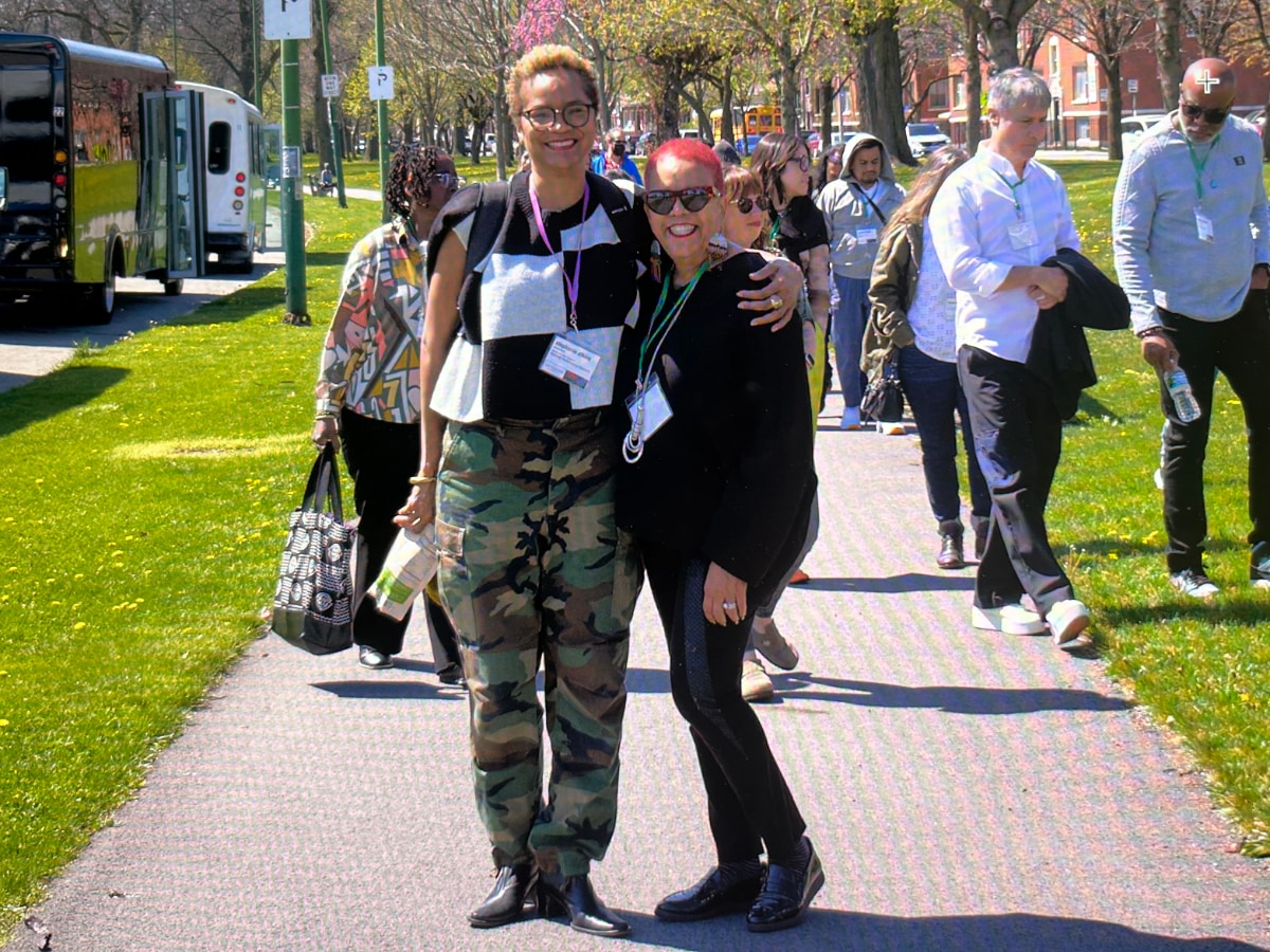 Stephanie Atkins and DVCAI founder Rosie Gordon-Wallace, two Black women, pose together for a photo outside on a sidewalk. Behind them, other members of their group walk across the sidewalk to an unseen destination.