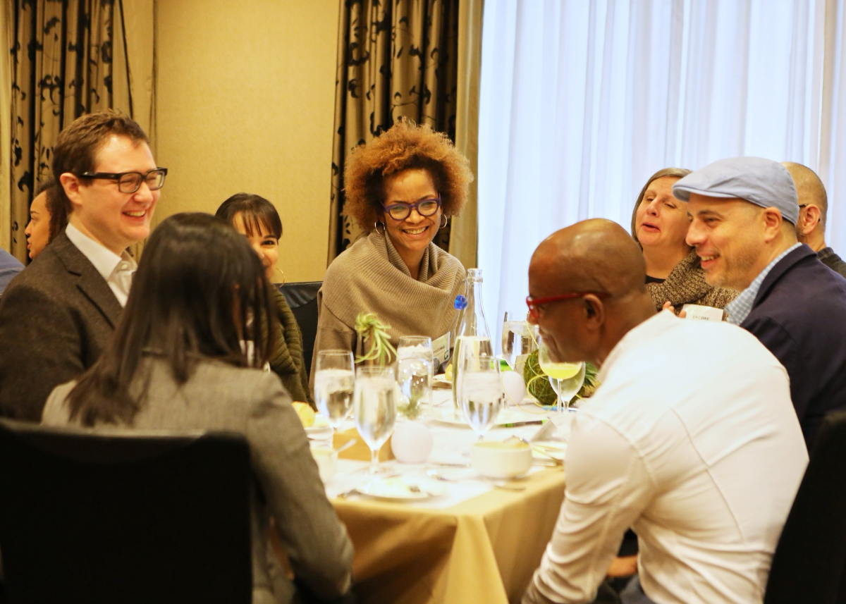 Stephanie Atkins, a Black woman, sits at a cloth covered dining table surrounded by men and women of various ethnicities. The dinner guests are smiling and laughing as they chat with each other. There are large, stemmed glasses filled with iced water on the table, as well as place settings. The photo is framed so that Stephanie is the only person seen from the front, with the other guests shown in profile or from behind.