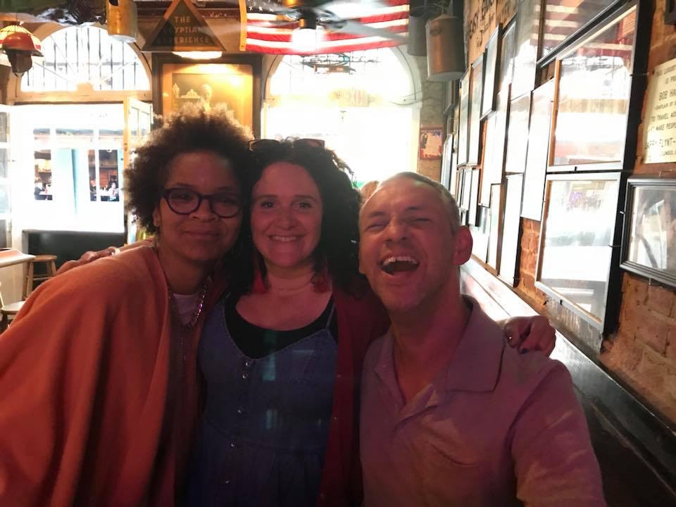 Three people pose together indoors, likely at a restaurant or bar, with framed photos covering one wall and the American flag hanging from the ceiling. The windows behind them open to a brightly lit street, and the bright sunlight blows out the highlights in the photo and leaves the three people in shadow. On the left is Stephanie Atkins, wearing glasses and an orange wrap. In the middle is a woman with dark, wavy hair and wearing a dark top under a blue denim dress. She is smiling and has her arms around the shoulders of the other two figures. On the right, a man with short hair and wearing a light purple collared shirt is laughing with his mouth open.
