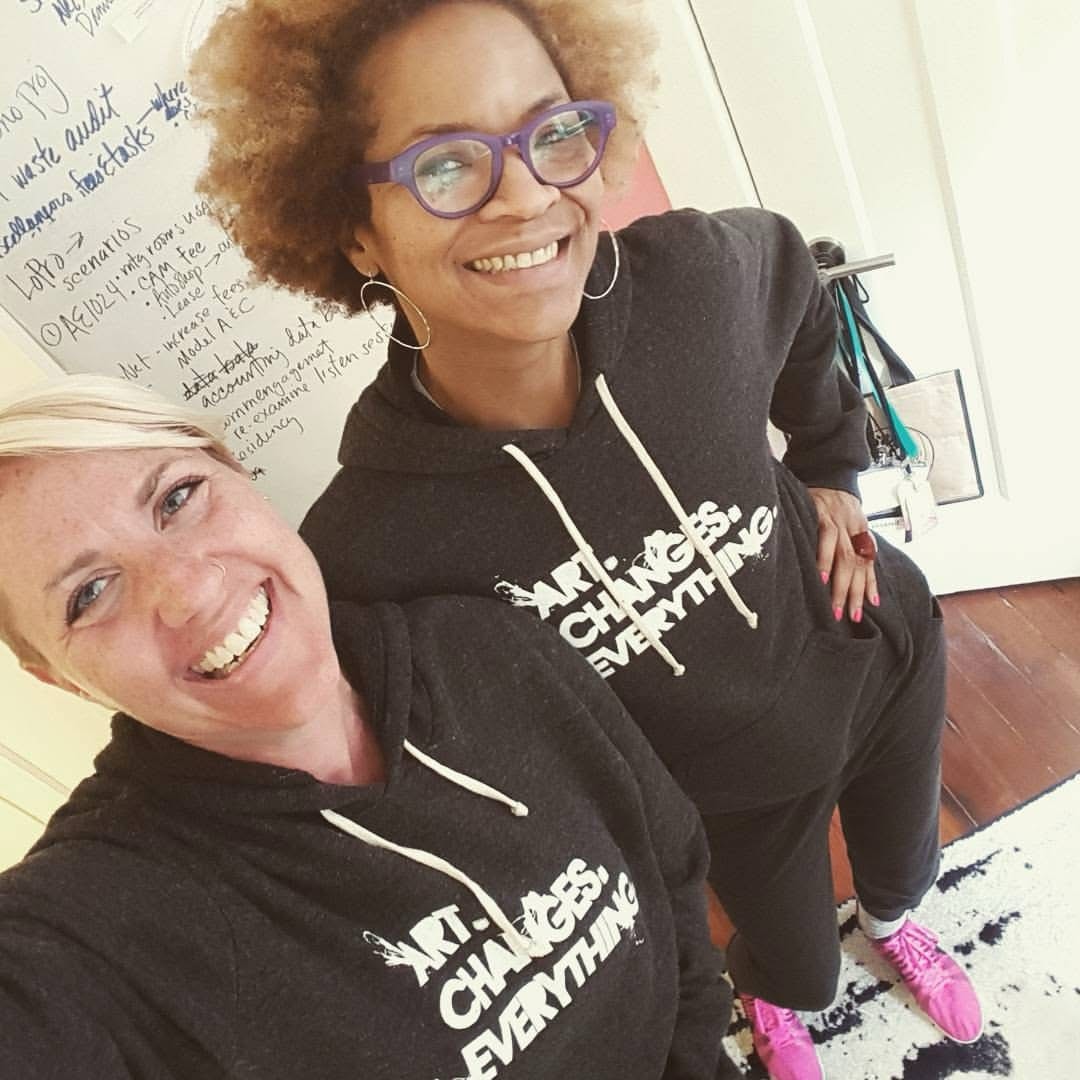 Stephanie Atkins stands with Caitlin Strokosch as they smile for a selfie. Each of them is wearing a black sweatshirt with the text "art changes everything" in white letters printed across the front. Caitlin is holding the camera above them so that their matching sweatshirts are fully visible.