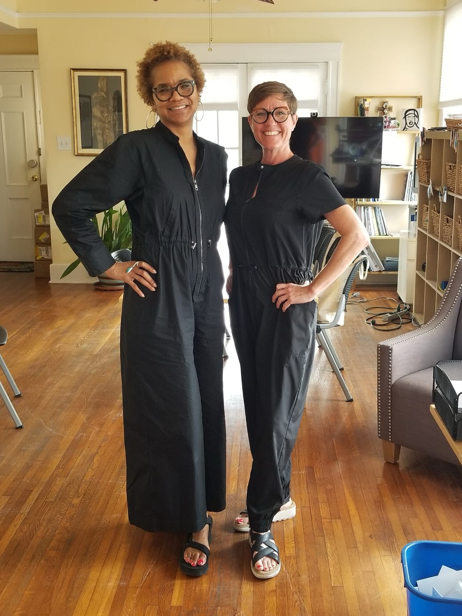 Stephanie Atkins stands next to Caitlin Strokosch in a full length photo that shows they are dressed in similar black one-piece suits that zip up the front, and sandals with black straps. The each have one hand on their hip and are turned slightly toward each other as they smile at the camera.