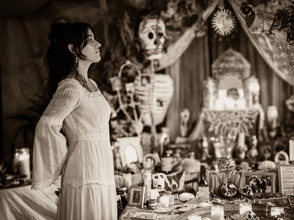 A photo is a side profile of a light-skinned woman standing before an elaborate Dia de Muertos altar lit with candles. She is a Chicana woman who wears a long-sleeved white dress; her hair is pulled back with loose curls surrounding her face. The image is a black-and-white print.