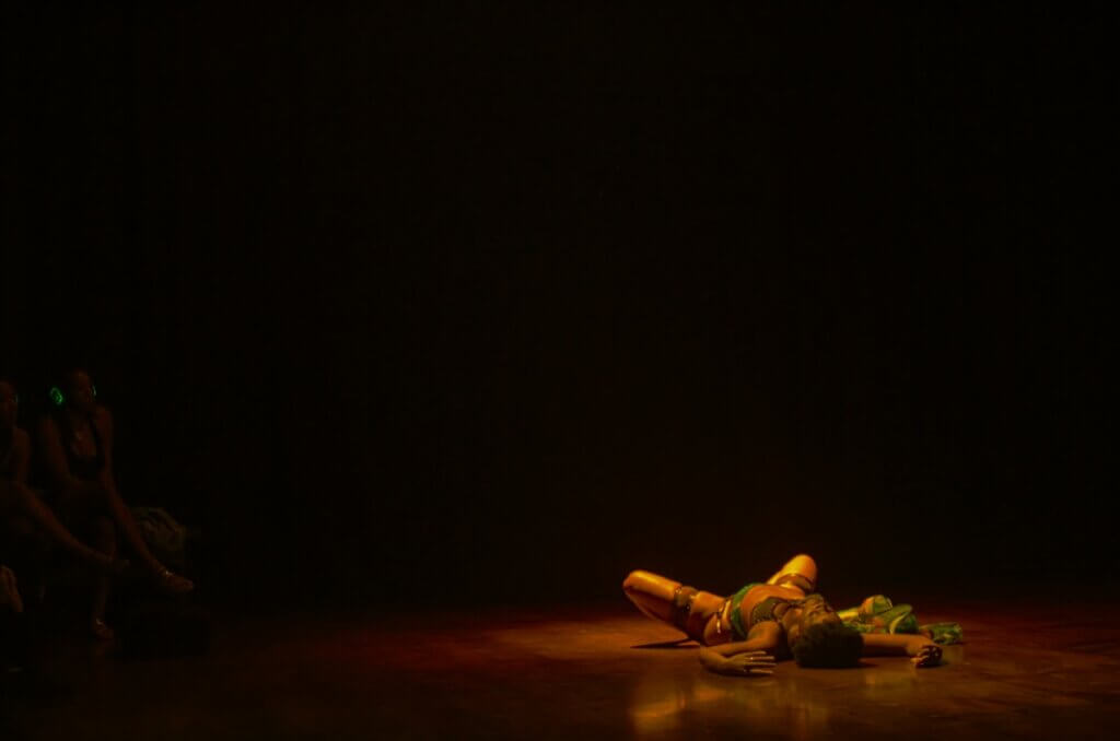 A brown-skinned woman lays on her back on a wooden dance floor, legs folded with feet touching and arms open above her head. An audience member can be seen watching with legs crossed, with silent disco headphones on and the green light glowing from the headphones.