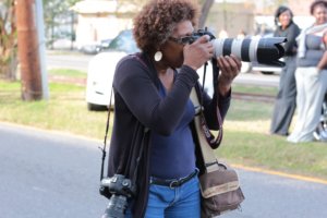 Chandra McCormick, a brown-skinned Black woman, brown hair with mingled gray, in her sixties, stands in the street with her camera in hand. She is wearing blue jeans with a navy blue cotton top, with a 35mm camera around her neck, another on her right shoulder. The camera has a long lens on it, and McCormick is poised to shoot the camera and make an image.