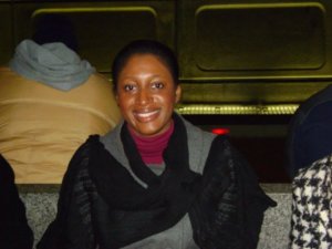 A dark-skinned woman is sitting on a subway bench smiling at the camera. It is late fall or early winter and she is wearing a sweater, coat, and shawl.