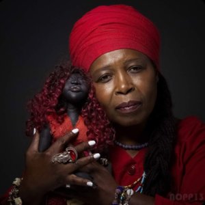 Rukiya, a brown skinned woman, wearing a red tignon and beaded necklaces, poses with a dark-skinned doll with red curls wearing a red dress.