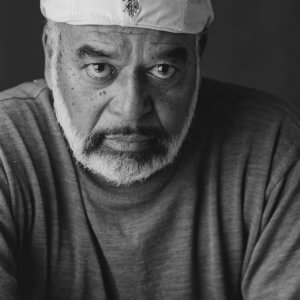 A black-and-white headshot of a Black man with a trimmed white beard. He wears a t-shirt and a white beret cap backwards and looks into the camera with a serious expression.