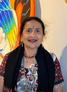 A medium-skinned woman with black hair, a bindi, and a nostril stud smiles. She is wearing a short-sleeved kurta with a burgundy floral pattern on it, two necklaces, and burgundy lipstick. Part of a brightly colored part of an artwork with a blue wing can be seen behind her.
