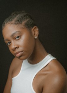 A medium-skinned woman posing for the camera from the breastline up in front of a black backdrop. Her hair twisted into six flat twists. She has big brown eyes, two earrings, and has a slight grin on her face. She is wearing a white muscle shirt and her head is tilted to the right.