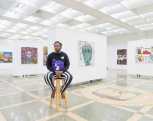 John Isiah Walton sits on a stool in a gallery wearing a graphic longsleeved shirt with a cartoon on it, black and white striped pants, and no shoes. He is surrounded by colorful, absurdist paintings on white walls.