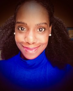 A close-up, bust-length view of a dark-brown skinned Black woman with shoulder-length, black, curly hair dressed in a royal blue turtleneck shirt. She is smiling with her mouth closed and facing the sunlight.