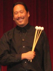 An Asian American man holding four marimba mallets smiling at the camera. He is wearing a black button-down shirt, a contrast to the white-yarn marimba mallets, against a background of a deep red theater curtain.