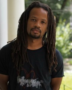 A medium skinned Black man with long locs and a black t-shirt standing on a porch.