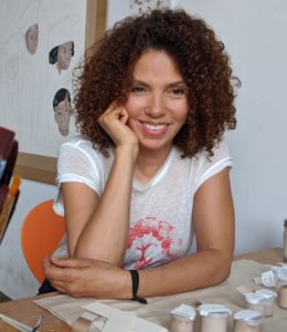 A middle-aged Black woman with medium light skin tone sitting at a desk, smiling at something outside of the camera. She has a dark brown shoulder length curly afro and she is resting her chin on her hand. Her elbow is propped up on the desk which has containers of paint on it. There is a painting of Black children in the background in muted tones.