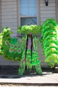 A New Orleans Masking Indian posing with his arms wide open in a green feathered multicolor rhinestoned suit. His wing is spread on the left side of the photo under his feathered arm and his war spear is on the right side of the photo in his hand. He wears a round feathered sun hat on his head with a multi green feathered war skirt around his waist. His eyes are fixed on his Big Chief awaiting a signal to move out and begin the journey across Mid-City.