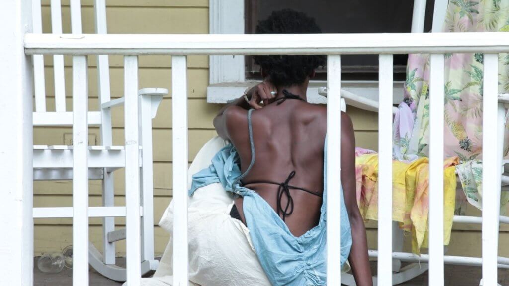 A photo of an African American woman sitting on a porch between two white rocking chairs with her back facing front, wearing a backless light blue top and white linen bottoms. With one arm bent resting her elbow on one knee, the other arm is straight with her hand laying flat on the porch.