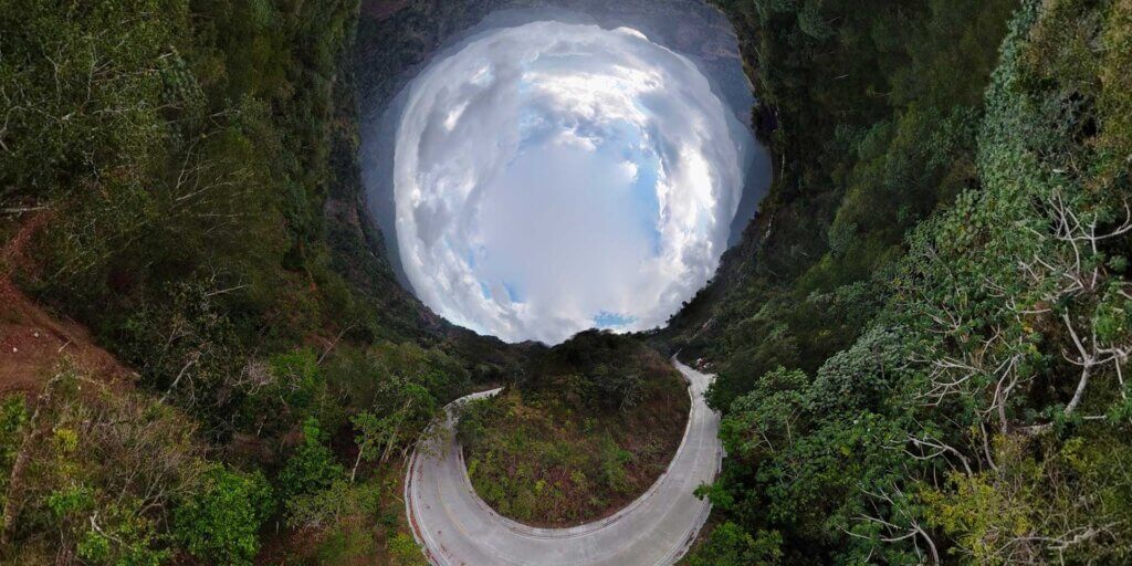 A daytime image of mountains in the distance using a 360 camera has trees in the foreground wrapping around the perimeter of the image with the sky forming a circle in the upper center. In the lower center of the image, a road curves down and back up, mirroring the circle of sky above.