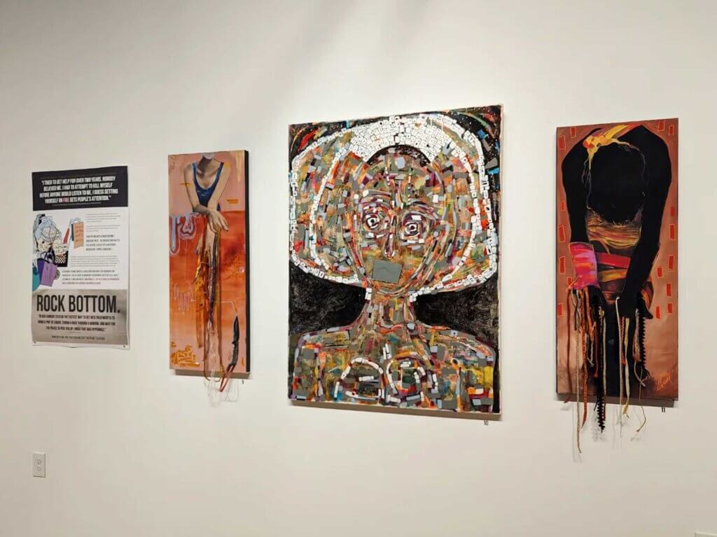 Four pieces of art are hanging on a white gallery wall; the first is a poster that says, "Rock Bottom" and features an illustrated image of a cart that holds an unsheltered individual's items and objects. Next to it are three paintings, one featuring a light-skinned individual in a blue tanktop from the shoulders down extending hands that have real orange and blue ribbons sewn to the canvas. The second is an abstract woman's face screaming, made up of a collage of gold and gray shapes. The third is a black slouching figure with similar ribbons extended off the canvas.
