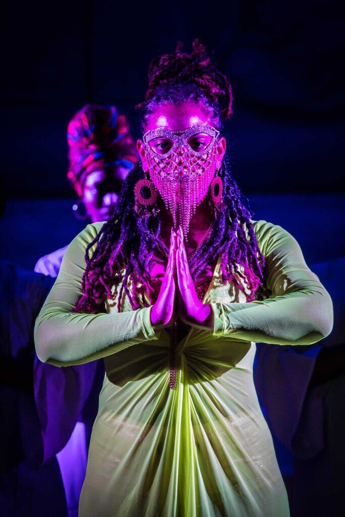A light-skinned Black woman with locks wearing a neon-green sheer dress and gold-sequined bra-let sings with her hands in a prayer pose.