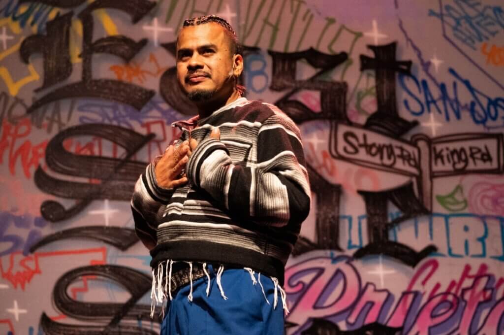 Close up of a medium-brown skinned man staring off into the distance, wearing a striped black and grey jacket, with blue pants, and in the background, a wall of graffiti.