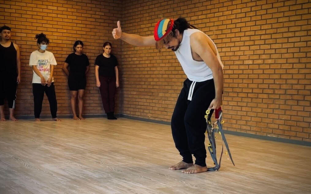 Brown-skinned Mexican American dancer Esteban Rosales wears a white sleeveless top, black pants, and a red, blue, yellow headpiece with brown fringe. Rosales stands barefoot in the center of a brick-walled room. His head is tilted down, back curved, knees bent, right arm shoulder height, palm facing inward and thumb up. His left hand is at his side, holding another headpiece decorated with gray ribbon and yellow flowers. On the side of the room, a group of dancers are lined up on the wall, with their focus on the singular dancer at the center of the room.