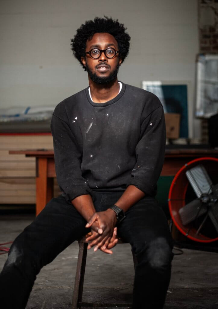 An image of a dark-skinned African American man wearing black-rimmed eyeglasses, a black sweatshirt, and black pants–looks at the camera while sitting on a stool in their studio.