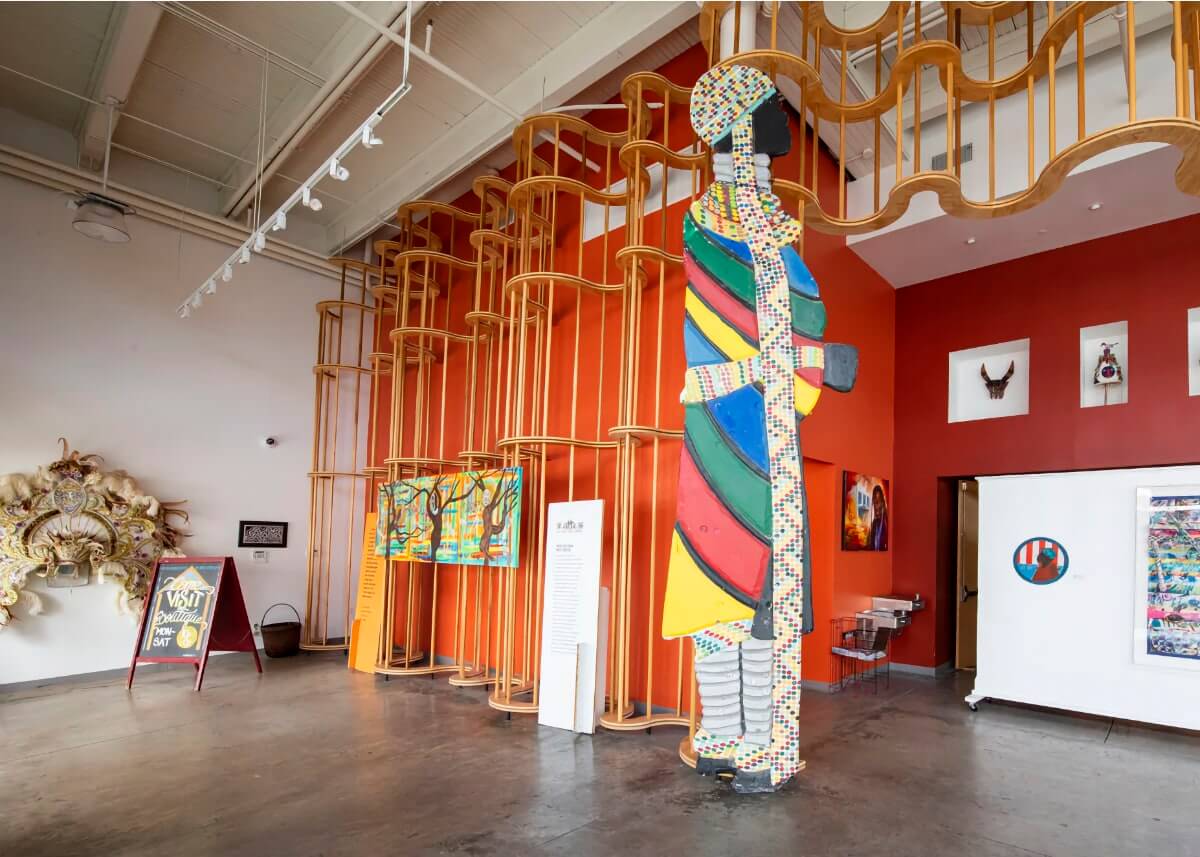 Interior of the Ashé Cultural Arts Center. The large space is 20 feet tall, with a white side wall upon which is affixed a five-foot-wide gold ornamental headdress, and a back wall that is painted a bold orange. Against this orange wall, a curving wooden scaffolding structure in blonde wood undulates the length of the wall. At the edge of the orange wall is a freestanding, 15 foot tall, two-dimensional representation of a Black person wearing a robe with yellow, red, green, and blue alternating stripes; and a belt, neck wrap, and cap with a floor-length ribbon all made from a white material with colorful beaded patterns.
