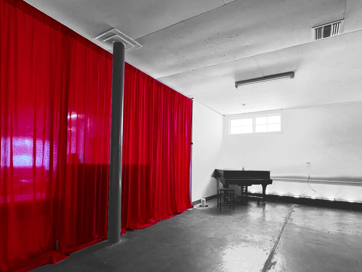 An empty performance space with white walls. A long red stage curtain runs nearly the full length of the wall on the left side, obstructed at the halfway point by a dark building support column. In the back corner, beneath small, bare windows along the top of the back wall, is a black grand piano with the top closed.