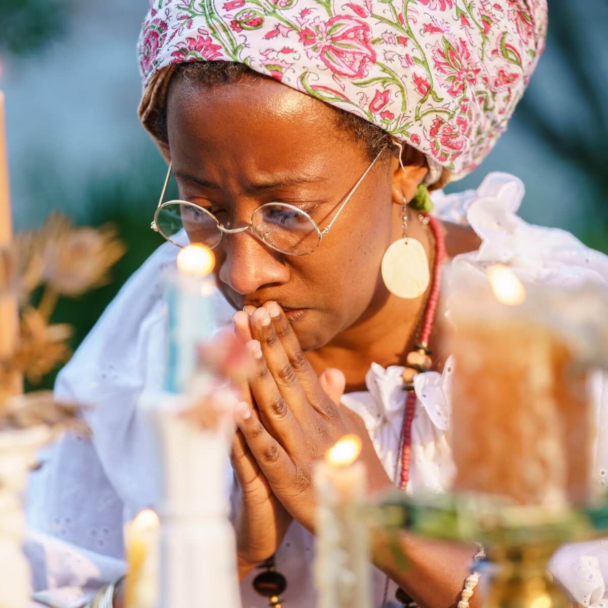 A medium-brown skinned woman in a tignon (head wrap) and wire rimmed glasses, praying at an altar made up of lit candles of different heights. The woman is clasping her hands flat together in prayer, eyes closes, and is wearing a white blouse, red necklace, and shiny round earrings.