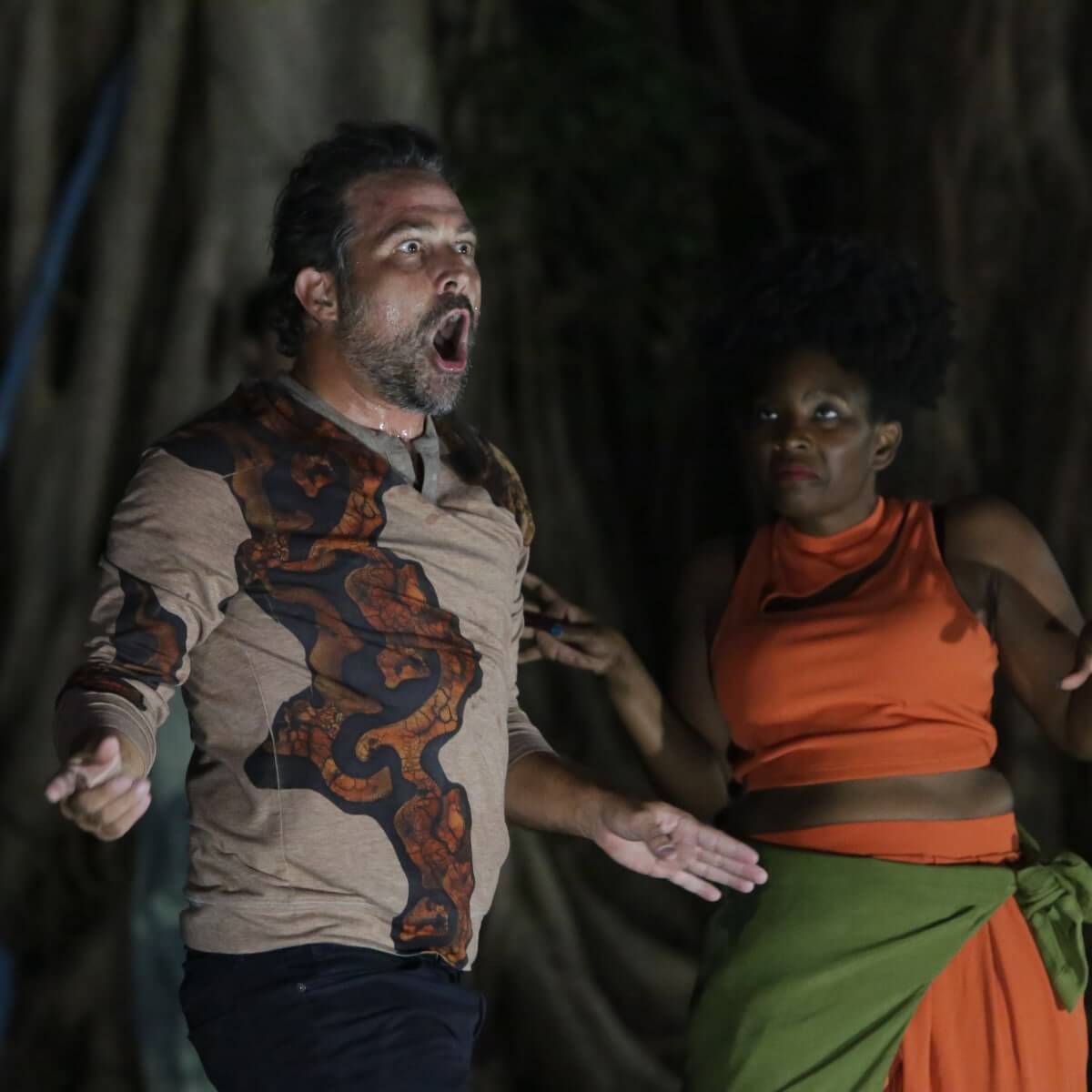 Two people are performing at night in front of a blurred background of trees. On the left, a man with a beard has his mouth open in an expressive pose, wearing a tan shirt with an ornate dark pattern featuring rust colored decorative elements. On the right, a Black woman wearing a bright orange dress with a green wrap tied around her waist watches him, her hands raised. They are illuminated from below by an off-camera light source.