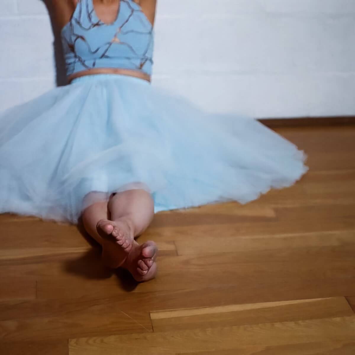 A tightly cropped photo of a female dancer sitting against the wall of a dance studio. Her arms and head are out of frame. she is wearing a light blue halter top adorned with an intersecting pattern of irregular, widely spaced bronze lines, and a light blue tulle ballerina skirt. Her legs are stretched out before her, with one bare foot resting on top of the other.