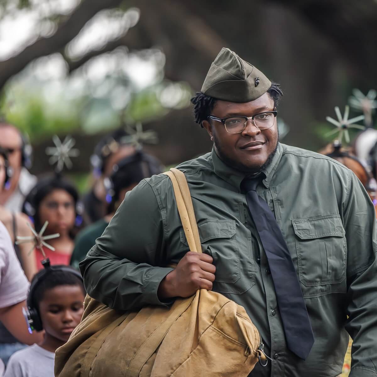 A Black man in military-style clothing smiles warmly as he walks past the camera. He's wearing glasses, a green military-style cap, a green button-up shirt with chest pockets, and a dark tie. He carries a large tan duffel bag over his shoulder. The photo is outdoors with a blurred background showing other people, suggesting this might be at some kind of public performance or processional.