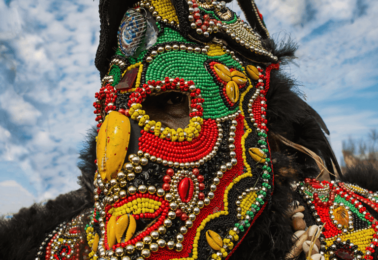A person wearing an elaborate traditional ceremonial mask and costume adorned with intricate beadwork in vibrant colors including red, green, yellow, and black. The ornate headdress features detailed beaded patterns, metallic elements, feathers, shells, and other decorative materials against a blue sky with white clouds. The craftsmanship displays complex geometric and organic designs typical of indigenous ceremonial regalia.