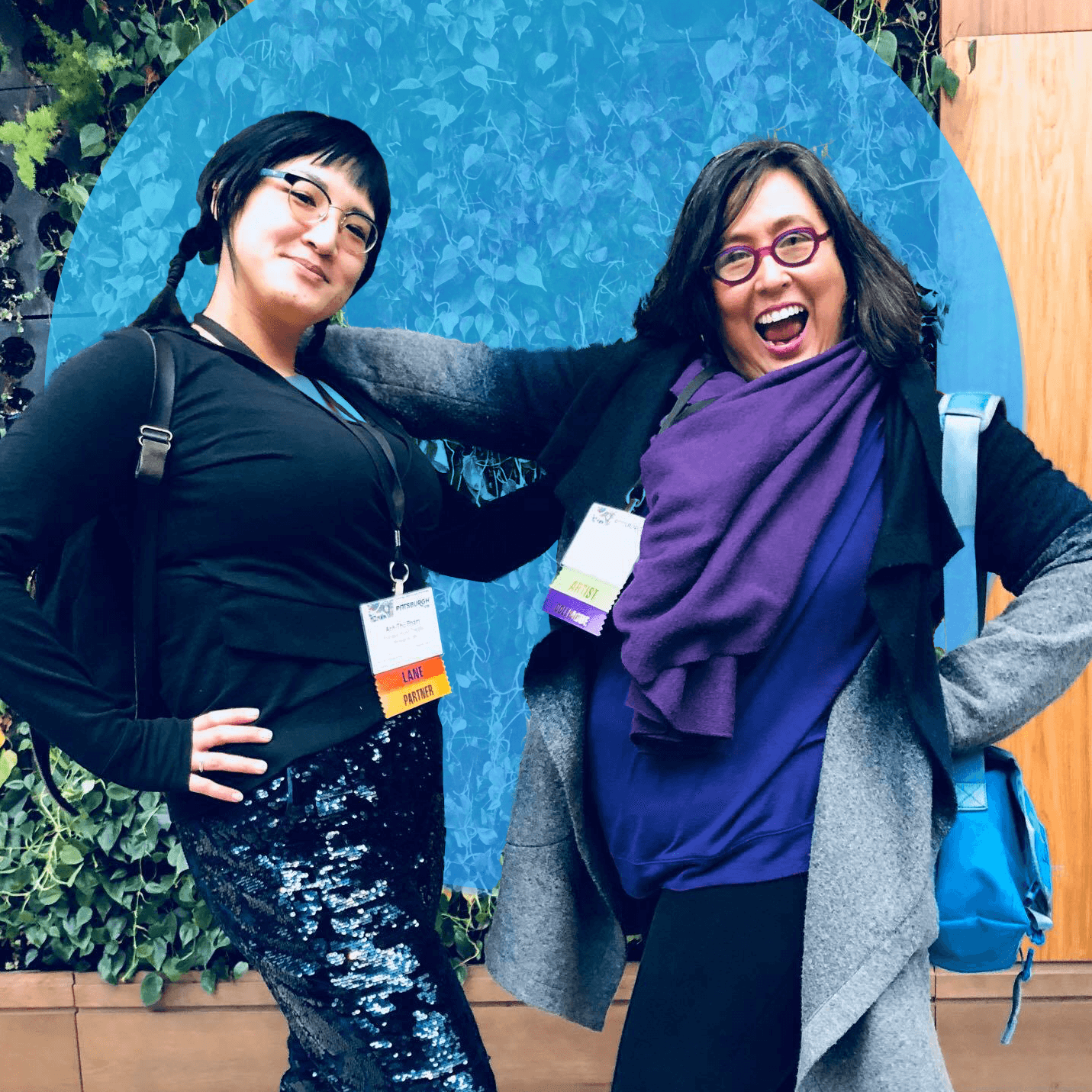 Two women pose playfully in front of a lush green plant wall. The woman on the left wears glasses, a black top, sparkly dark blue sequin pants, and black boots, while the woman on the right, also wearing glasses, sports a bright purple scarf, grey cardigan, dark pants, and reddish-purple shoes, smiling widely with a hand on her hip and a turquoise backpack over her shoulder. Both wear conference badges around their necks.