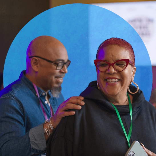 A smiling woman with short reddish hair, cat-eye glasses, large hoop earrings, and a black hoodie stands holding a smartphone, while a man in glasses and a denim shirt gently places his hands on her shoulders from behind. In the blurred background, several people sit working at tables, with a large screen and a whiteboard visible, suggesting a conference or workshop setting.