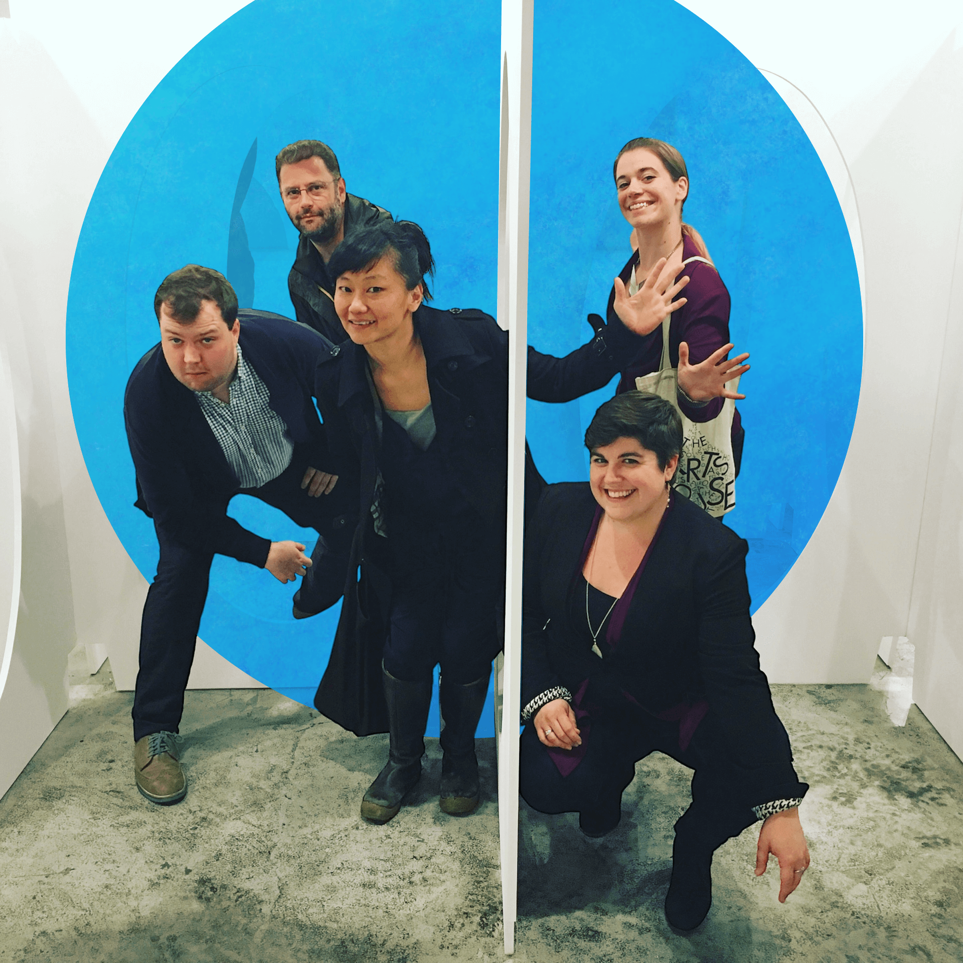 Five people pose playfully among large white sculptural panels with oval cutouts. Three people stand or crouch on the left side of a partition, leaning through the openings, while two others mirror them on the right, smiling and gesturing with their arms extended. The group appears cheerful and engaged in an interactive art or design installation space with a concrete floor.