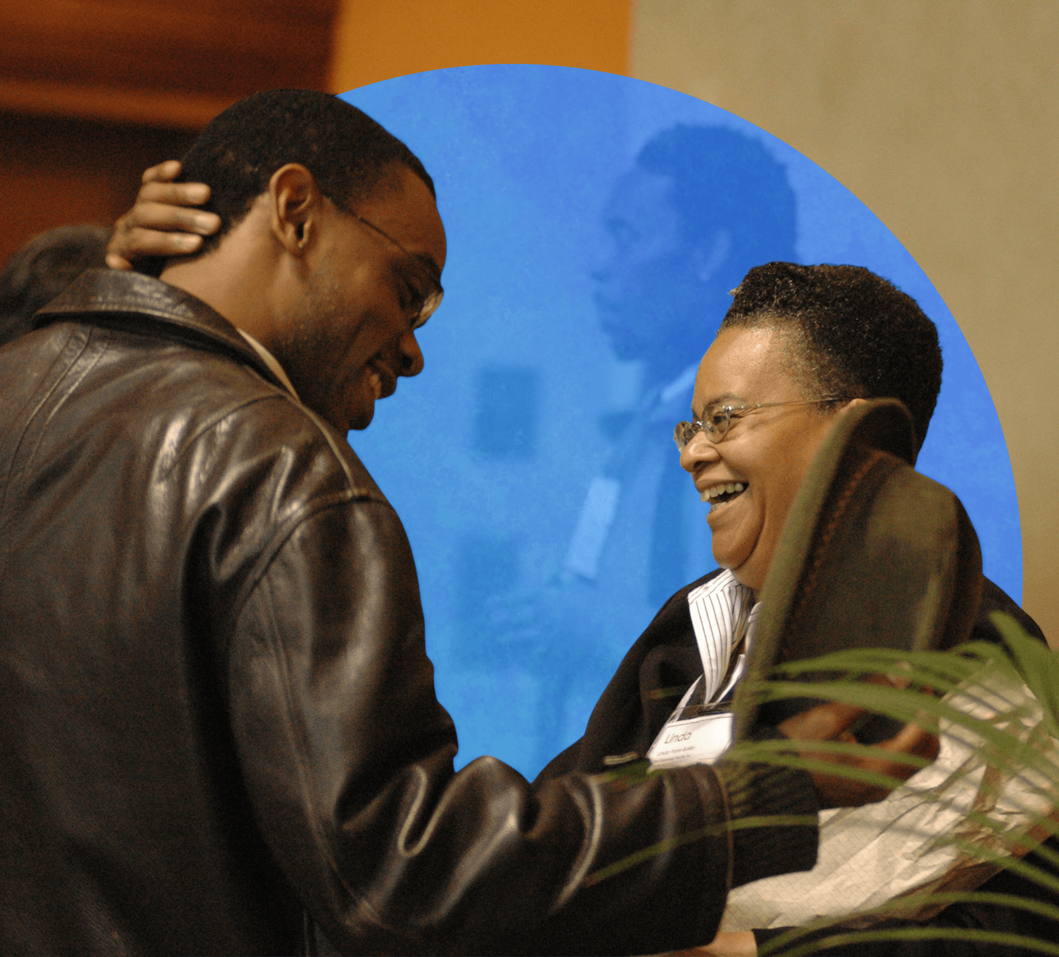 A man in a brown leather jacket smiles warmly while greeting a woman holding a hat, who is laughing joyfully. Both wear glasses and conference name badges, standing in an indoor setting with warm lighting, while another man in a grey sweater and scarf appears slightly blurred in the background. A green plant leaf is partially visible in the foreground.