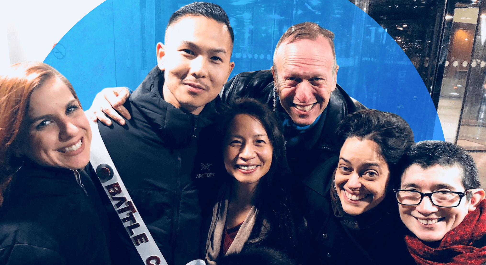Six people stand closely together outdoors at night, smiling brightly for the camera. One person wears a white sash reading “BATTLE CHAMPION” over a dark jacket, holding a cup, while the others, bundled in coats and scarves, lean in around him. Glass doors and city lights reflect in the background, giving a festive, urban vibe.