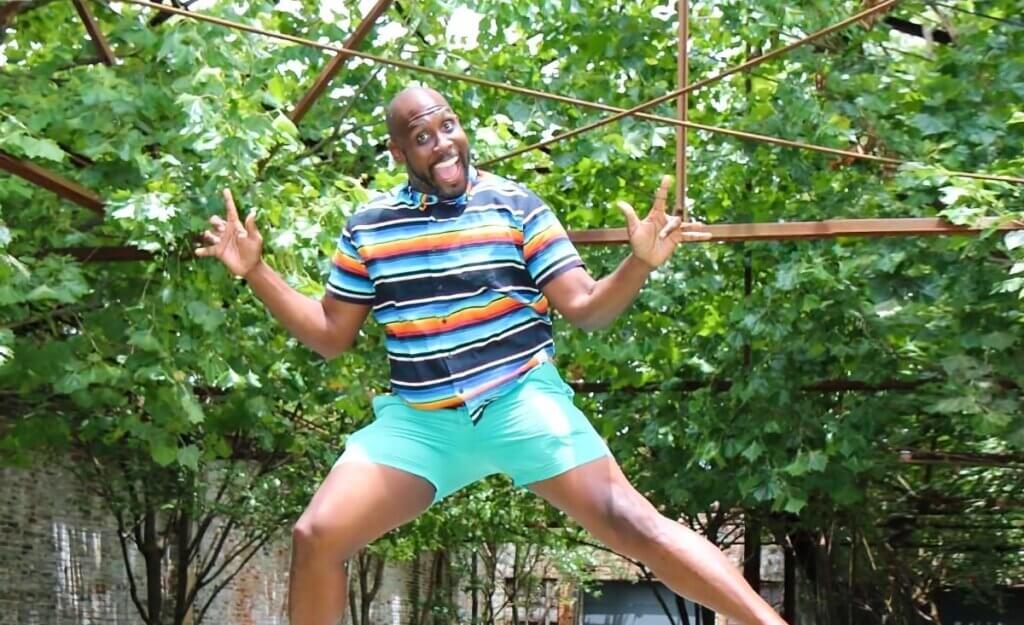 An outdoor, enclosed patio space, with brick walls and a small table and two chairs in the background. In the foreground, a young Black man with a bald head and a dark, close cropped beard is smiling at the camera as he jumps into the air. He is wearing a buttoned shirt with alternating horizontal stripes of dark blue, white, light blue, and orange, and he's wearing aqua colored shorts and pink lace-up sneakers. Above and behind him is a canopy of leaves from the shade-providing trees scattered around the patio.