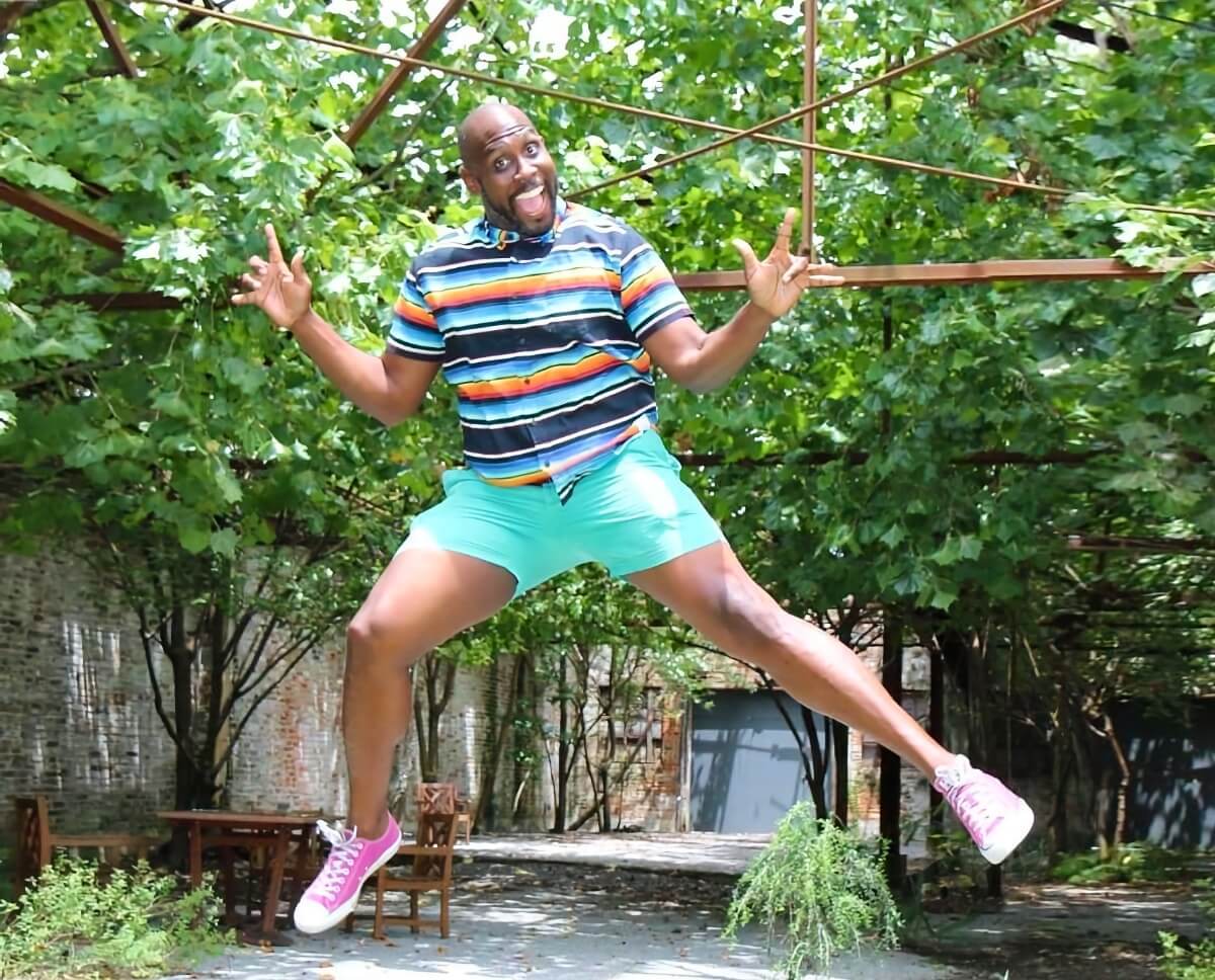 An outdoor, enclosed patio space, with brick walls and a small table and two chairs in the background. In the foreground, a young Black man with a bald head and a dark, close cropped beard is smiling at the camera as he jumps into the air. He is wearing a buttoned shirt with alternating horizontal stripes of dark blue, white, light blue, and orange, and he's wearing aqua colored shorts and pink lace-up sneakers. Above and behind him is a canopy of leaves from the shade-providing trees scattered around the patio.