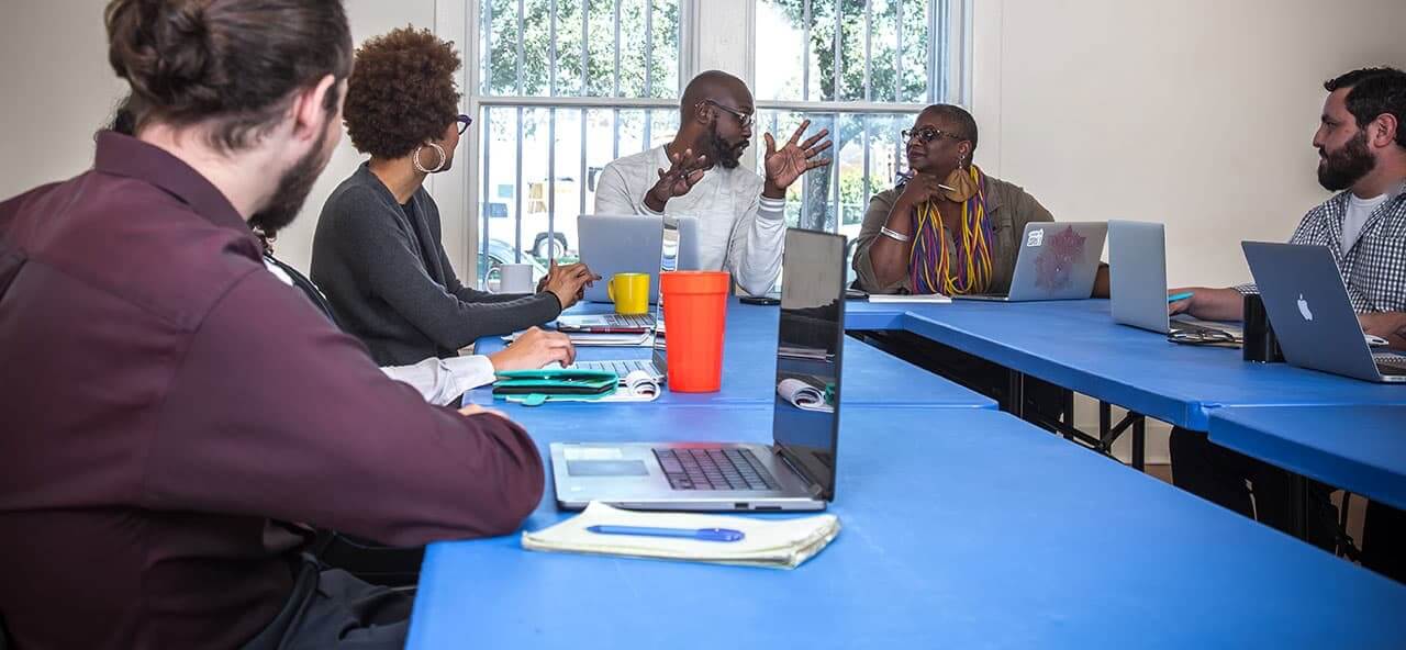 A conference room with blank white walls. Five long blue tables are pushed together in a U shape to enable a small group of NPN staff members to hold a meeting. In the center of the photo, at the end of the U shape, a Black man with a bald head, closely cropped dark beard, and glasses gestures with both hands as he speaks to a Black woman with closely cropped hair and a colorful scarf seated to his left. On either side of the table, the other four attendees sit with opened laptops and listen attentively. Large windows behind the group let in natural light, revealing greenery outside. The mood is serious and collaborative, with everyone focused on the discussion.