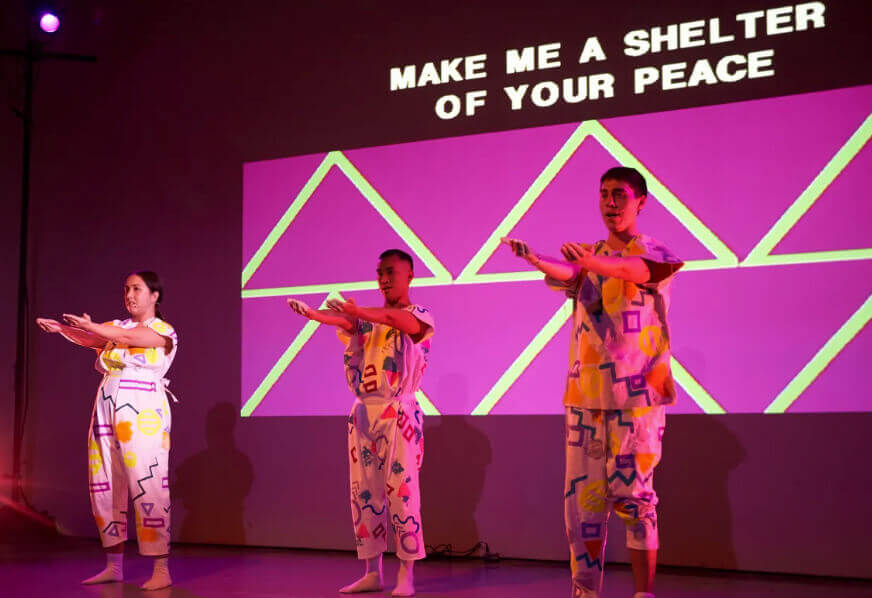 Three performers stand with their arms outstretched towards the audience. They are singing. They wear loose-fitting jumpsuits with geometric shapes printed on them and white socks. The first performer is a medium light-skinned woman with long dark hair. The next performer is a medium dark-skinned Filipino man with short dark hair. The third performer is a light-skinned man with short dark hair. They bask in the pink stage light, with a bright pink video projection behind them. The projection features a pattern of green triangles and the words “MAKE ME A SHELTER OF YOUR PEACE.”