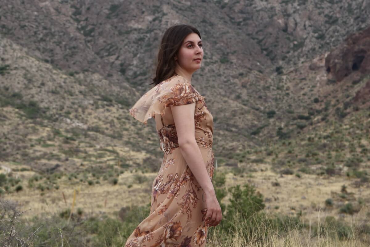 The artist Amanda Ekery, wearing a brown dress that blows in the wind, stands among the foothills in a southwest Texas landscape, with dry scrub brush around her.