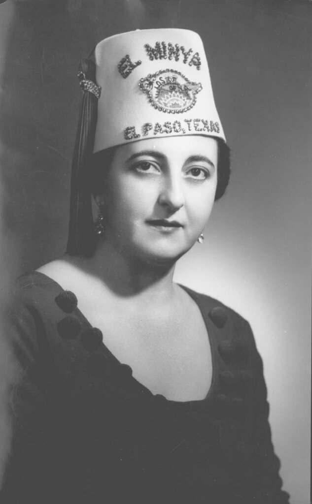 Black-and-white portrait of a woman wearing a fez hat that reads “El Minya, El Paso, Texas,” adorned with sequins and tassels. She has a calm, confident expression, and is dressed in a dark top with decorative fabric buttons. This image is noted to be Amanda Ekery’s great-sito Olga.