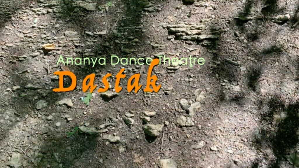 Text over a rocky dirt ground reads Ananya Dance Theatre in light green and Dastak in bold orange script. Small stones and patches of sunlight create a natural textured background beneath the title. The image serves as an opening title card introducing the dance work Dastak.