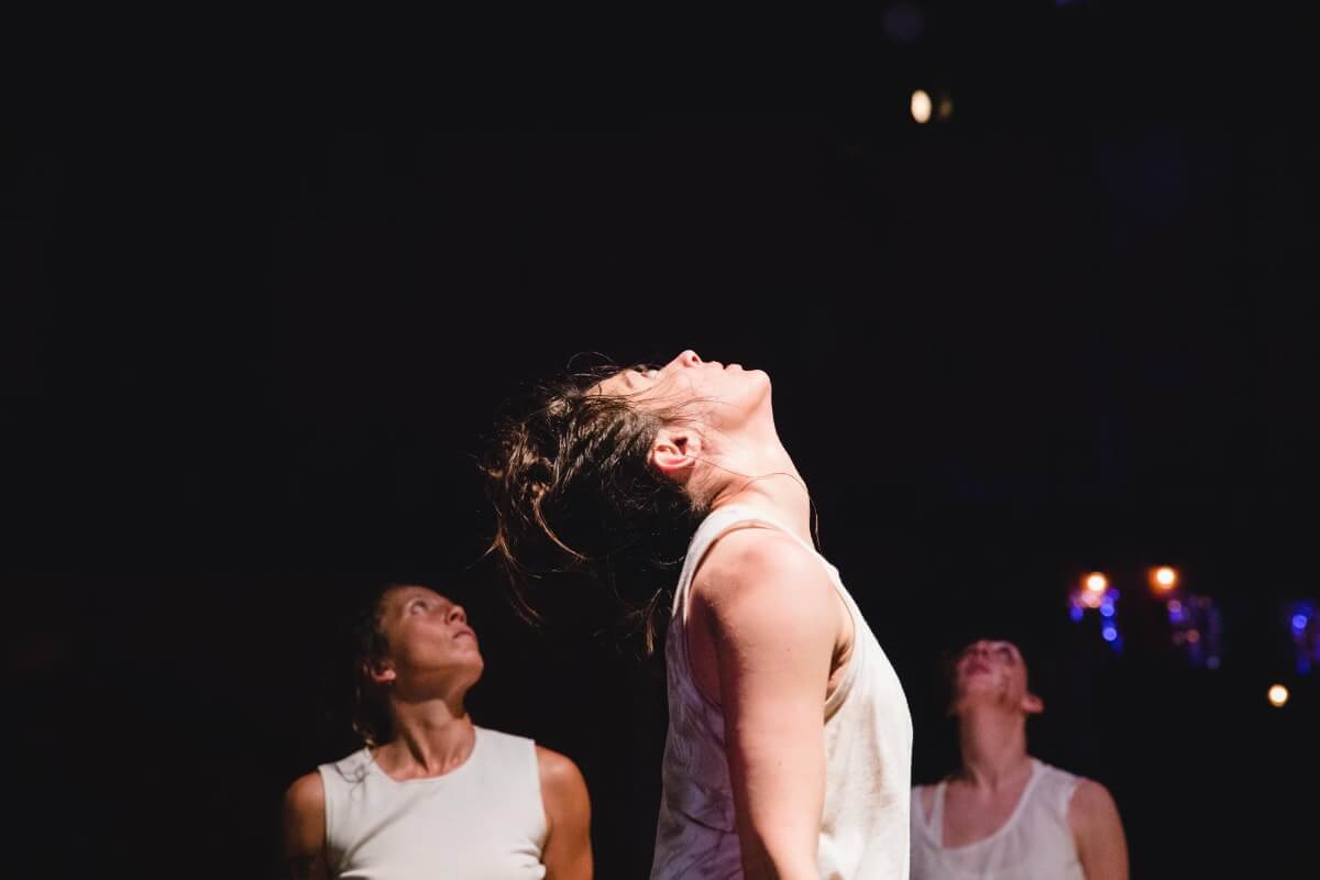 Three dancers in white stand on a dark stage and stare up.